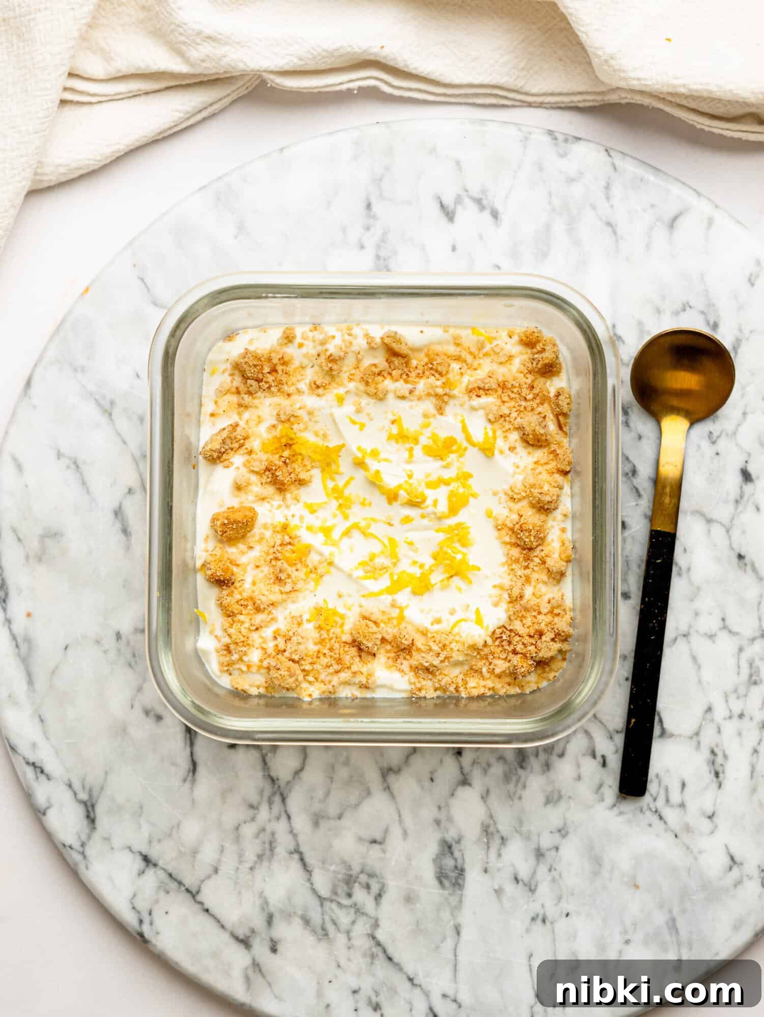 Lemon cheesecake overnight Weet-Bix in a glass container topped with biscuit crumbs and lemon zest, served on a marble board with a spoon alongside.