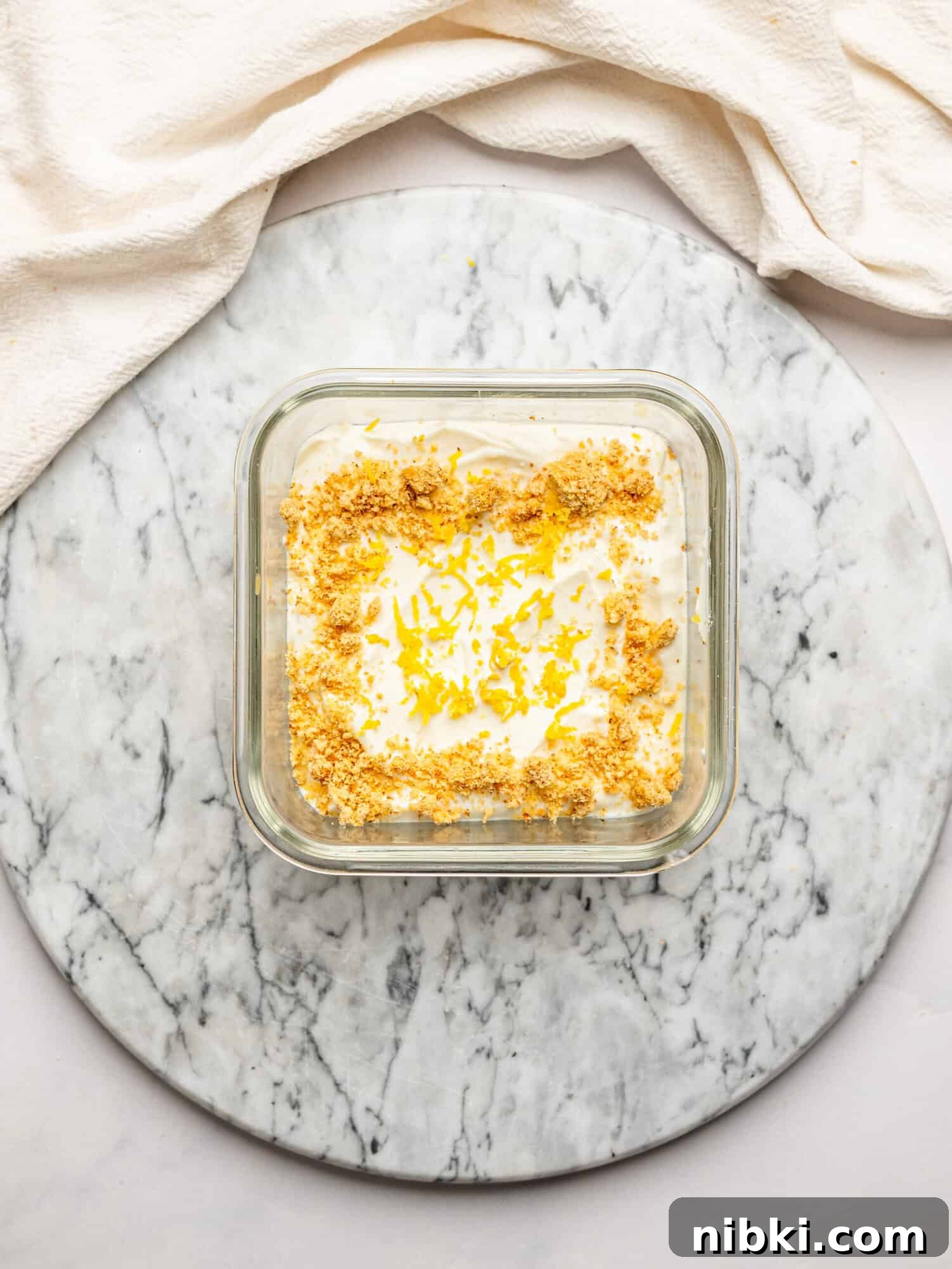 Overhead view of lemon cheesecake overnight Weet-Bix topped with biscuit crumbs and lemon zest, presented on a marble board with a draped cloth background.