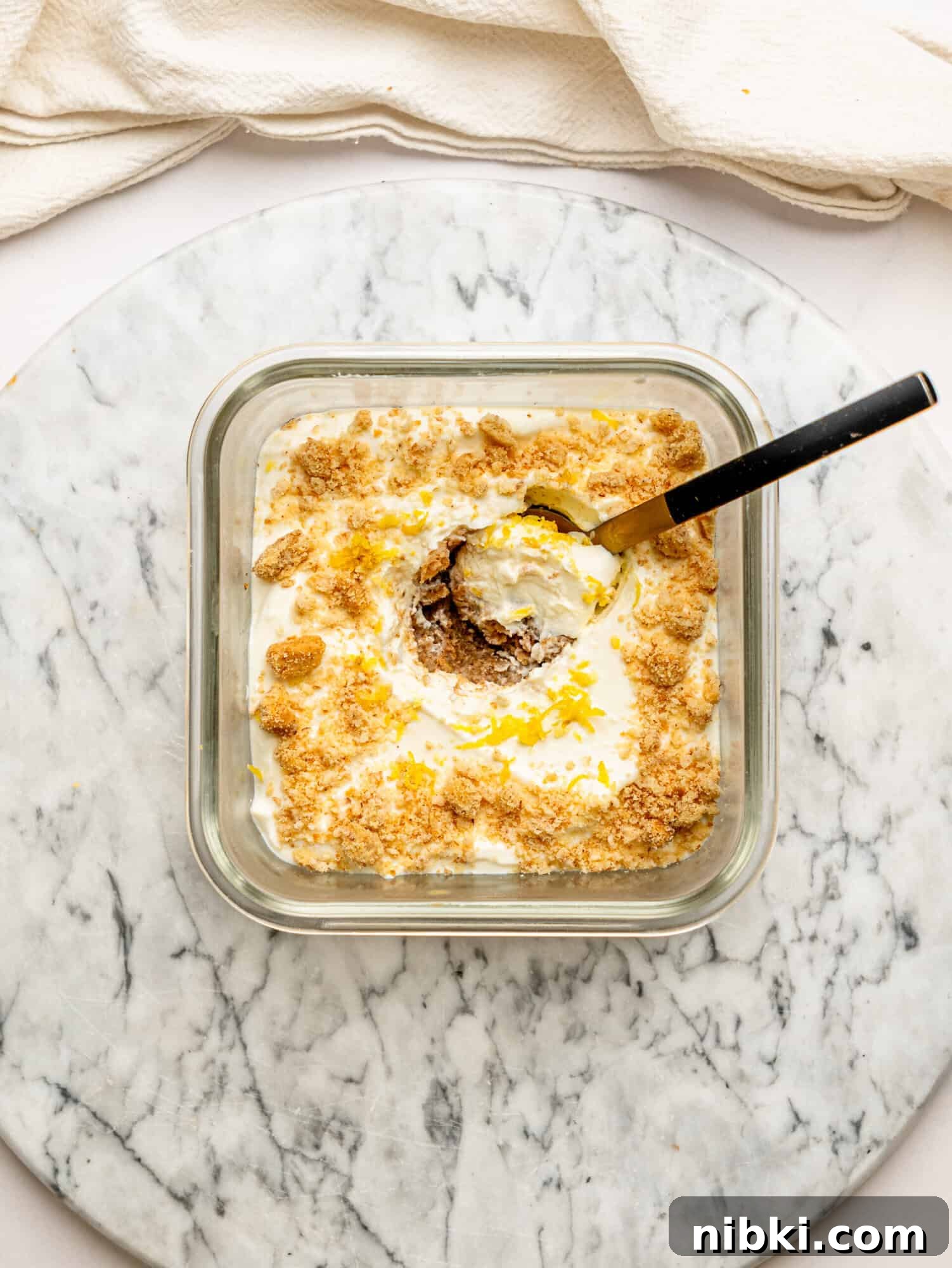 Lemon cheesecake overnight Weet-Bix in a glass meal prep container topped with crushed biscuit and fresh lemon zest, with a spoon scooping out a creamy serving.