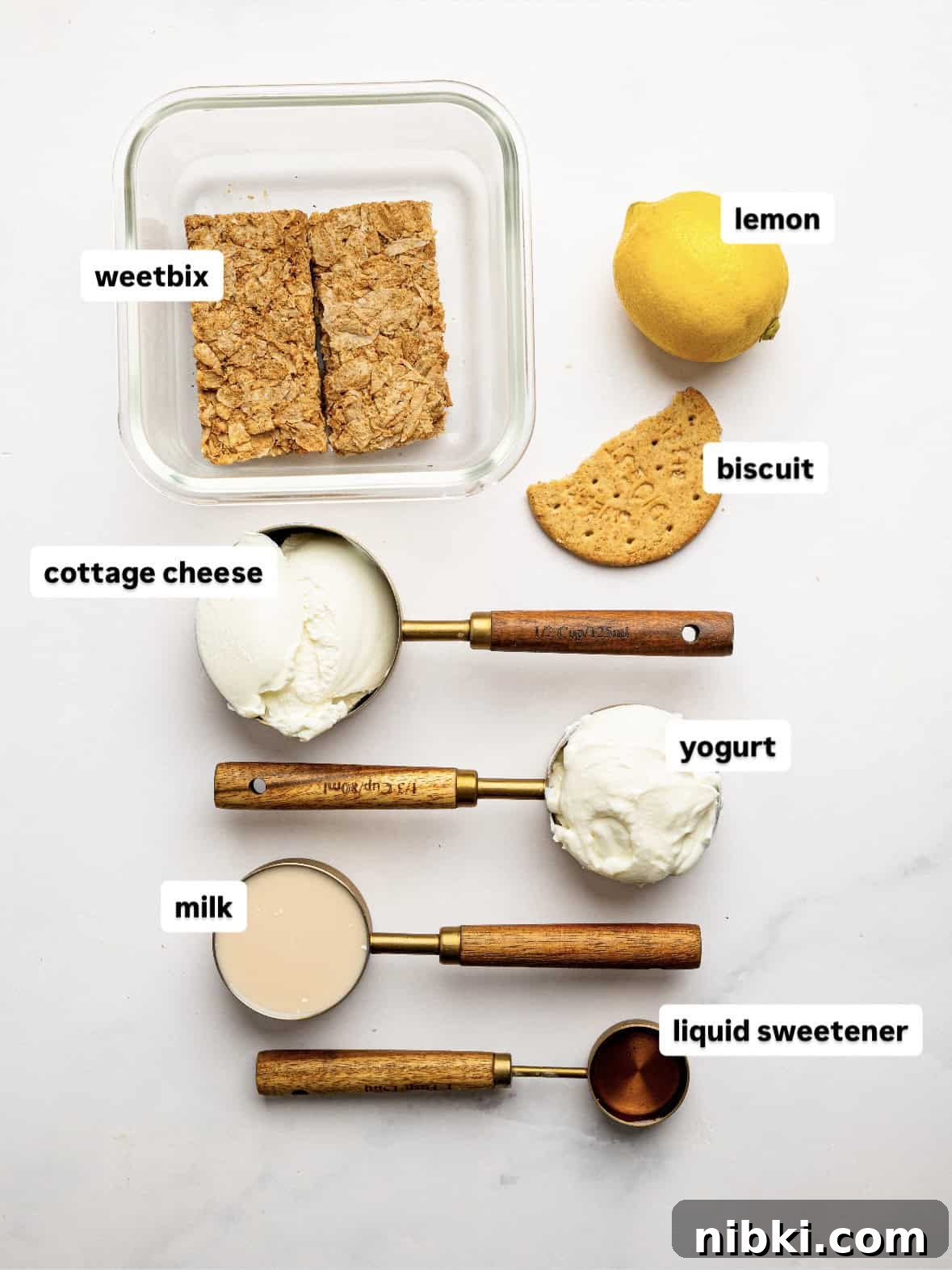 Ingredients for lemon cheesecake overnight Weet-Bix arranged on a white surface, including Weet-Bix biscuits, cottage cheese, Greek yogurt, milk, liquid sweetener, a lemon, and a biscuit for topping.