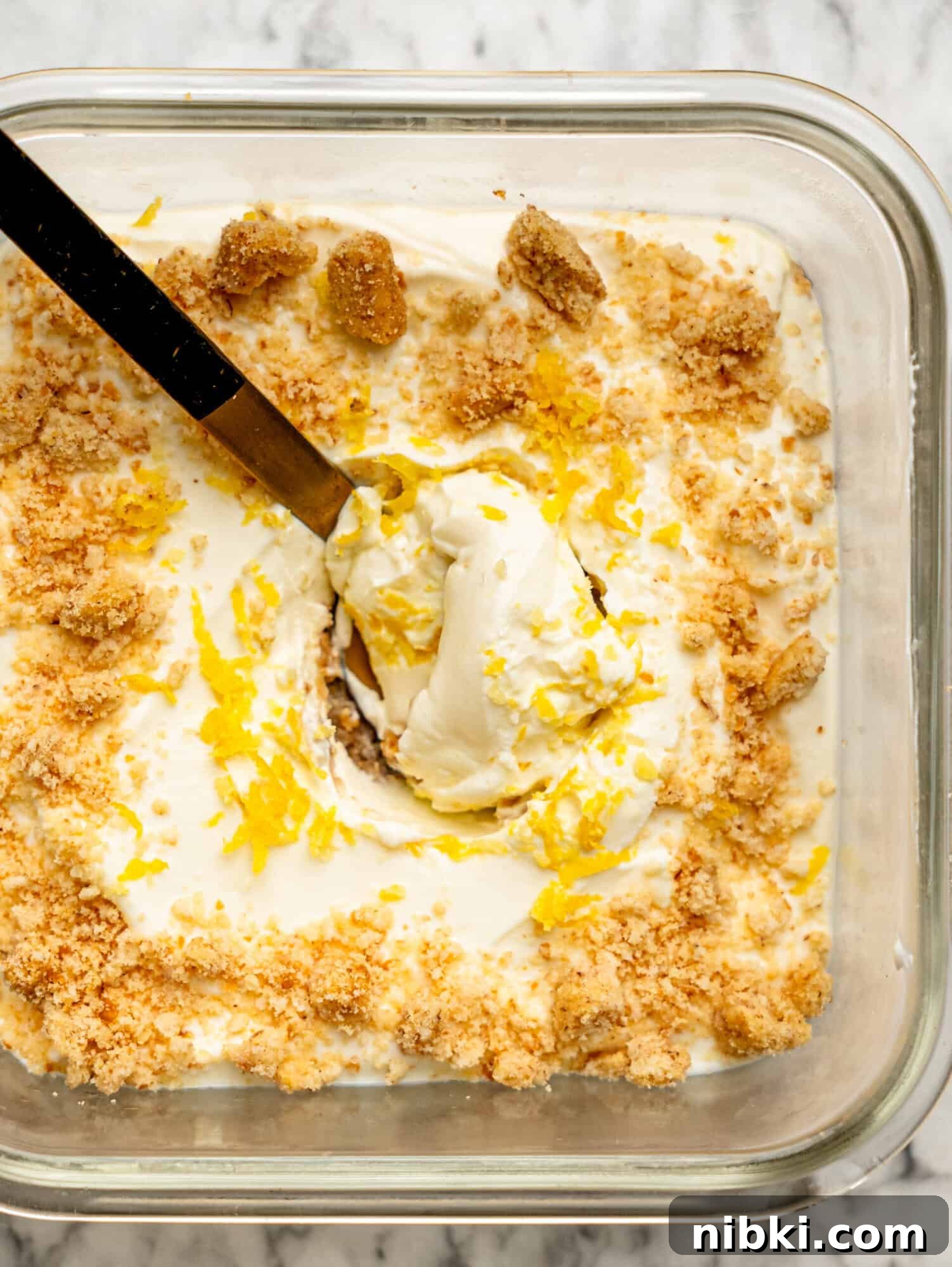 Close-up overhead view of lemon cheesecake overnight Weet-Bix with a spoon lifting the creamy lemon cheesecake layer, finished with biscuit crumbs and lemon zest.