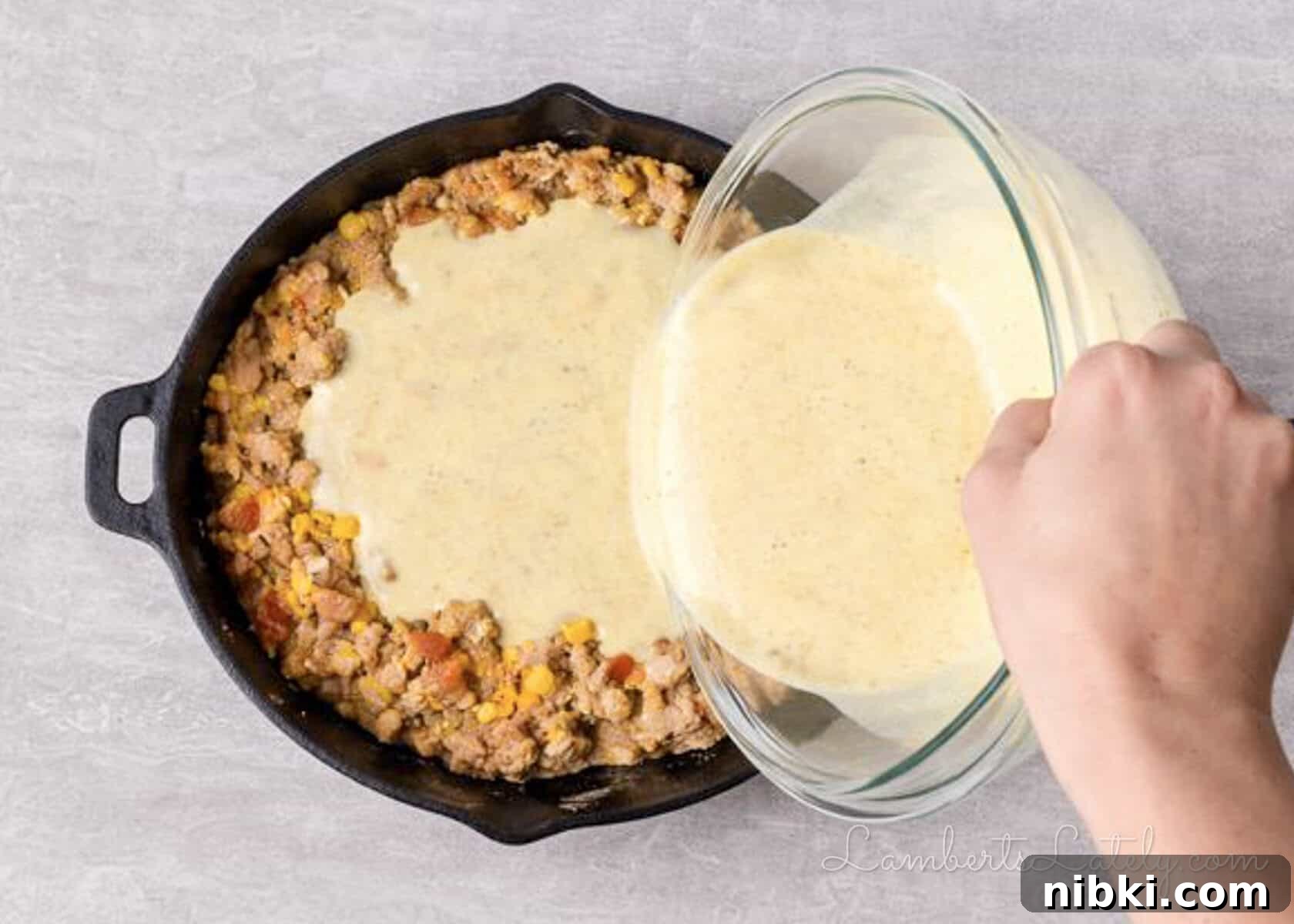 pouring cornbread batter over cowboy casserole mixture in a cast iron skillet.
