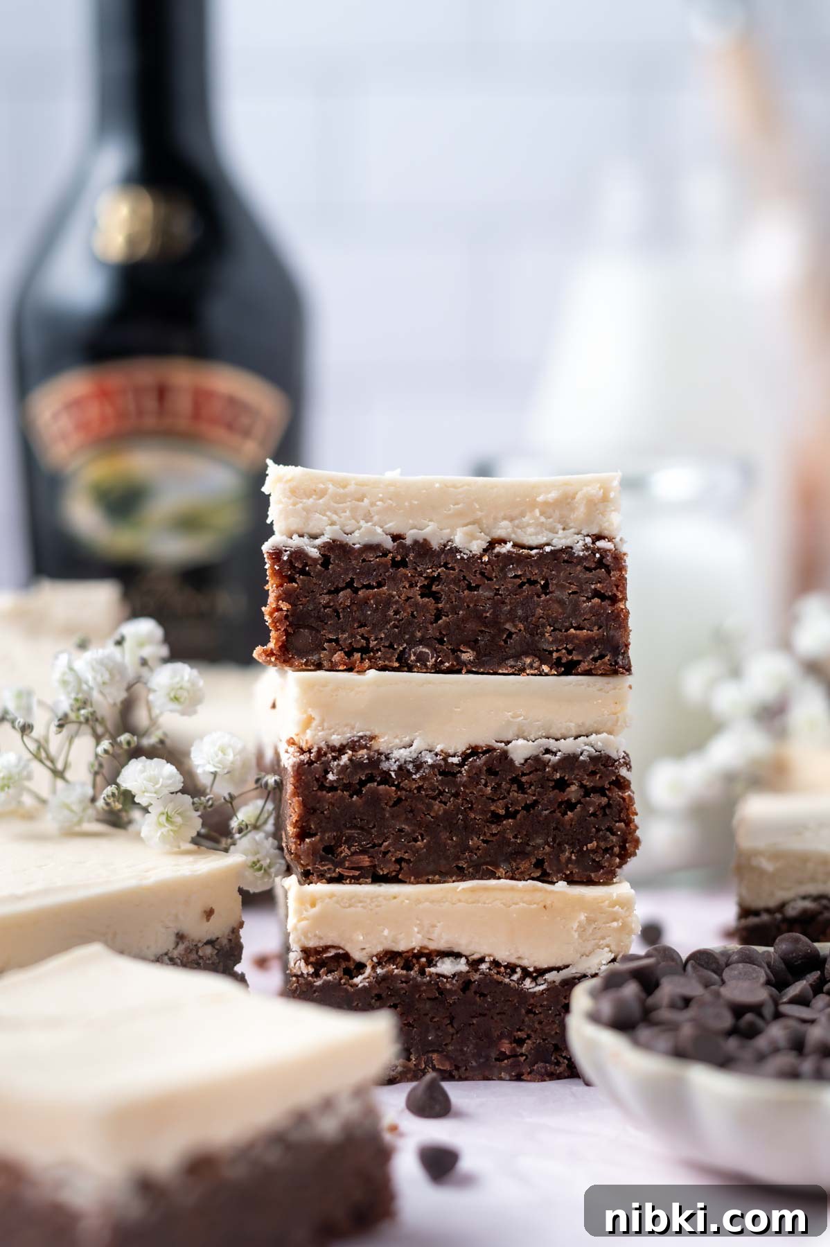 Fudgy Irish Cream Brownies Recipe 3 stack of frosted brownies with a bowl of chocolate chips in the foreground and milk in the background