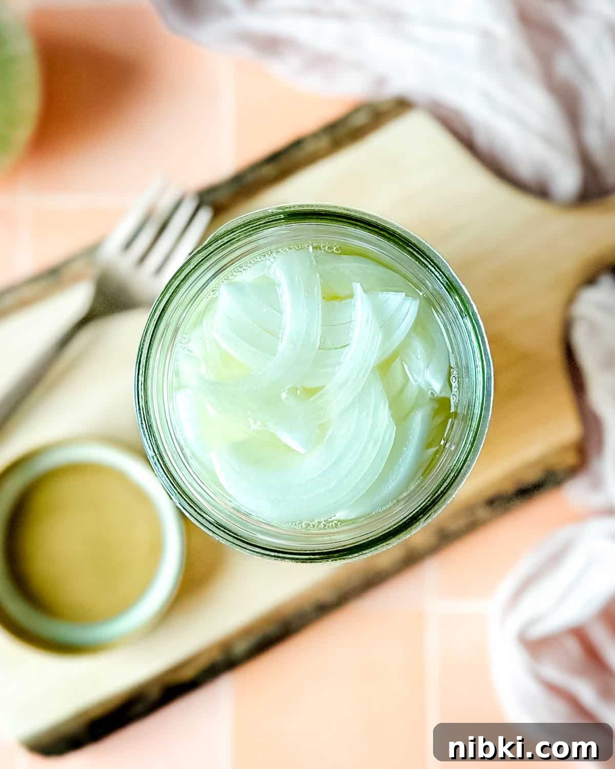 Quick Pickled White Onions for Tacos, Sandwiches and Salads 2 Pickled white onions in a glass jar on a wooden cutting board surrounded by white onions, a fork, and a pink linen.