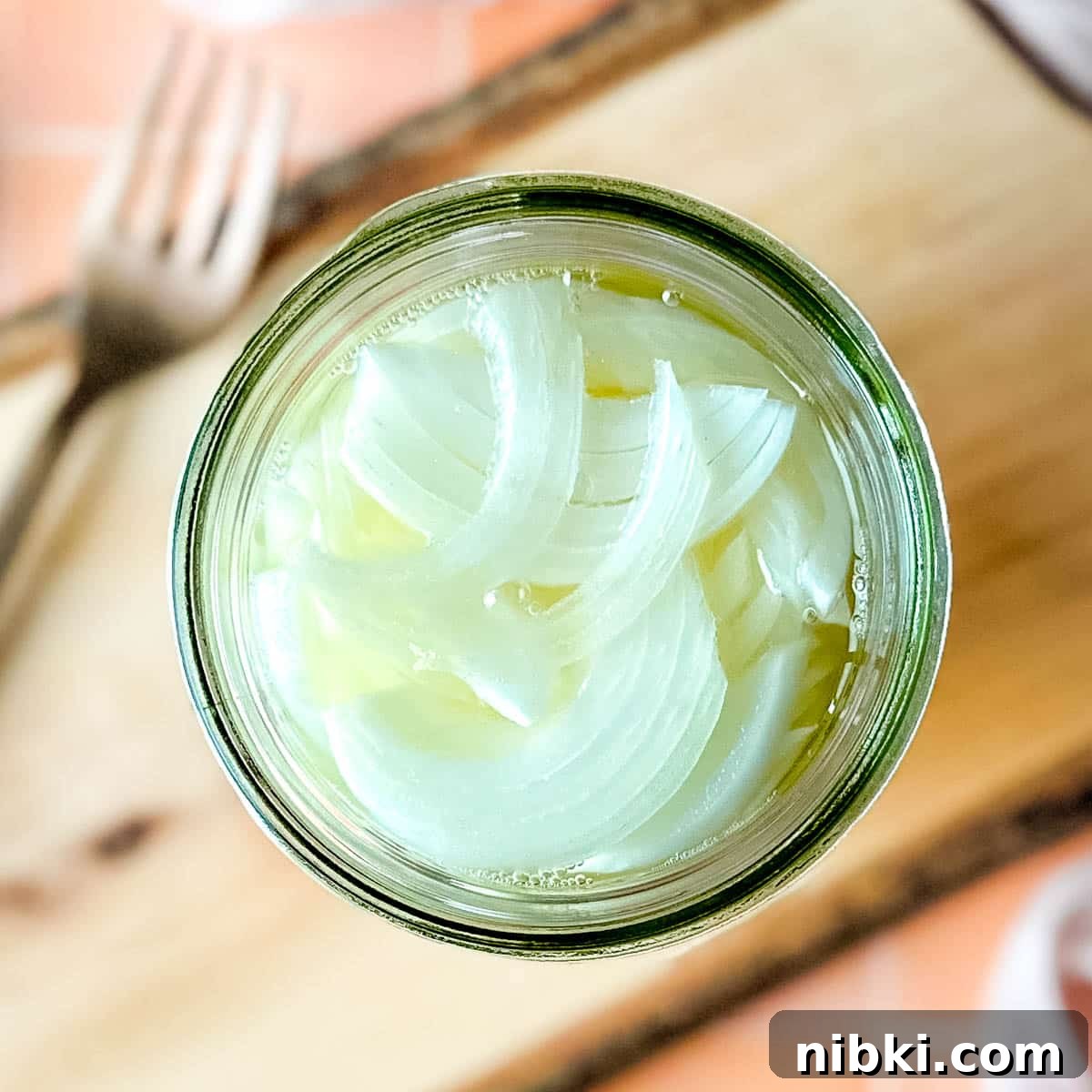 Quick Pickled White Onions for Tacos, Sandwiches and Salads 7 Pickled white onions in a glass jar on a wooden cutting board surrounded by white onions, a fork, and a pink linen.