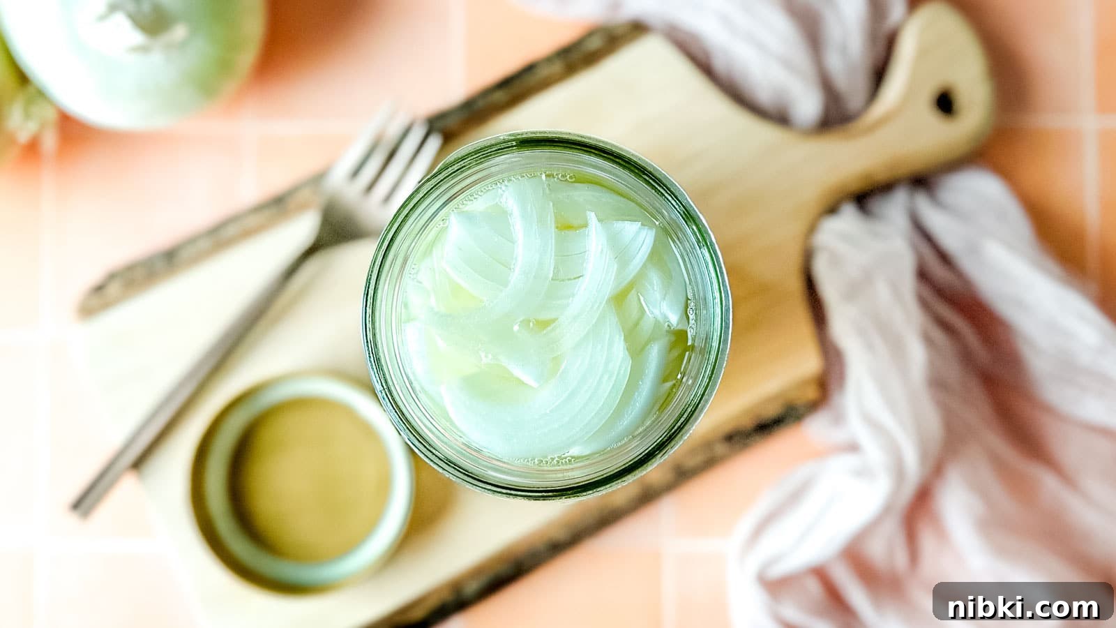 Quick Pickled White Onions for Tacos, Sandwiches and Salads 8 Pickled white onions in a glass jar on a wooden cutting board surrounded by white onions, a fork, and a pink linen.