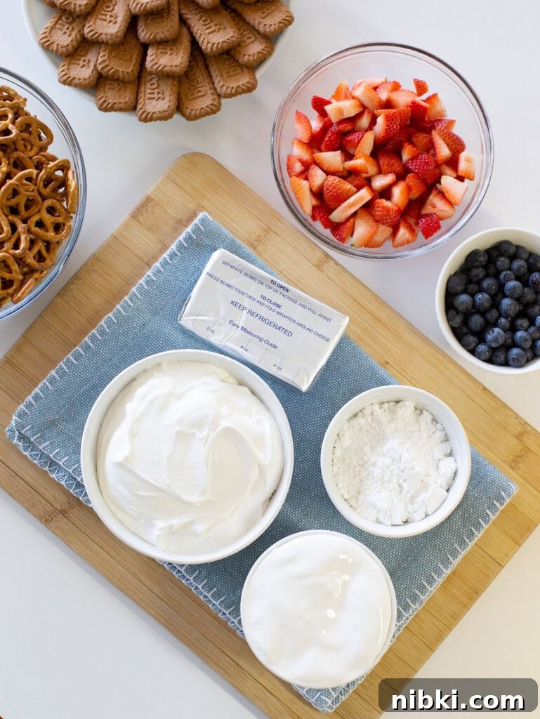 Patriotic Red, White and Blue Cheesecake Dip for 4th of July 3 A wooden board with bowls of whipped cream, powdered sugar, and cream cheese on a blue cloth. Nearby are dishes of sliced strawberries, blueberries, pretzels, and rectangular cookies.