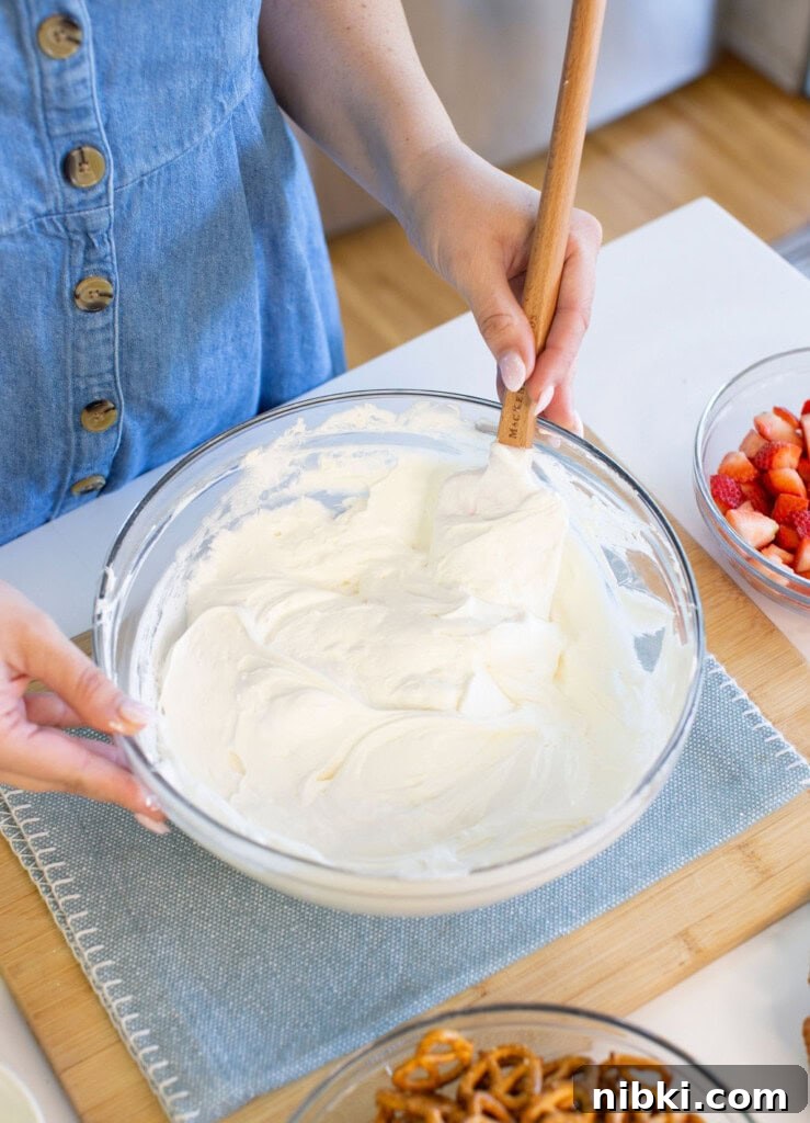 Patriotic Red, White and Blue Cheesecake Dip for 4th of July 4 Mixing whipped cream in a glass bowl with a spatula, bowls of sliced strawberries and pretzels nearby.