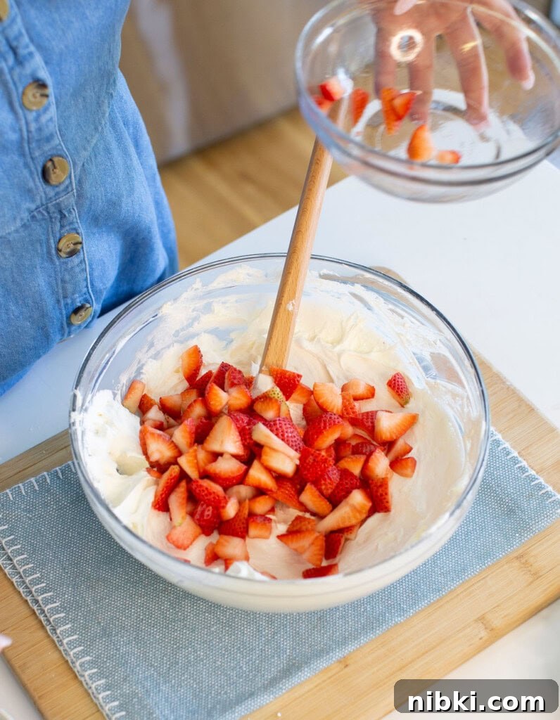 Patriotic Red, White and Blue Cheesecake Dip for 4th of July 5 Pouring chopped strawberries into a larger bowl filled with creamy batter and stirring with a wooden spoon.