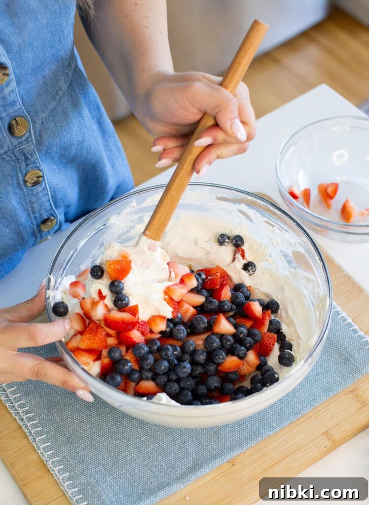 Patriotic Red, White and Blue Cheesecake Dip for 4th of July 6 Stirring a glass bowl of whipped cream, blueberries, and chopped strawberries on a wooden board.