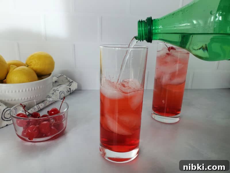 Sprite pouring into a red drink in a tall glass next to a bowl of maraschino cherries and a bowl of lemons.