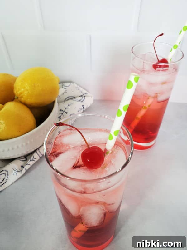 Looking down at two Dirty Shirley Drinks with maraschino cherry garnish and a paper straw next to a bowl of lemons.