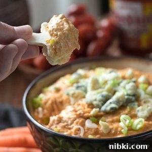 Low Carb Buffalo Chicken Dip topped with blue cheese and scallions in a brown bowl surrounded by vegetables. A piece of cauliflower is shown after being dipped into the hot appetizer.