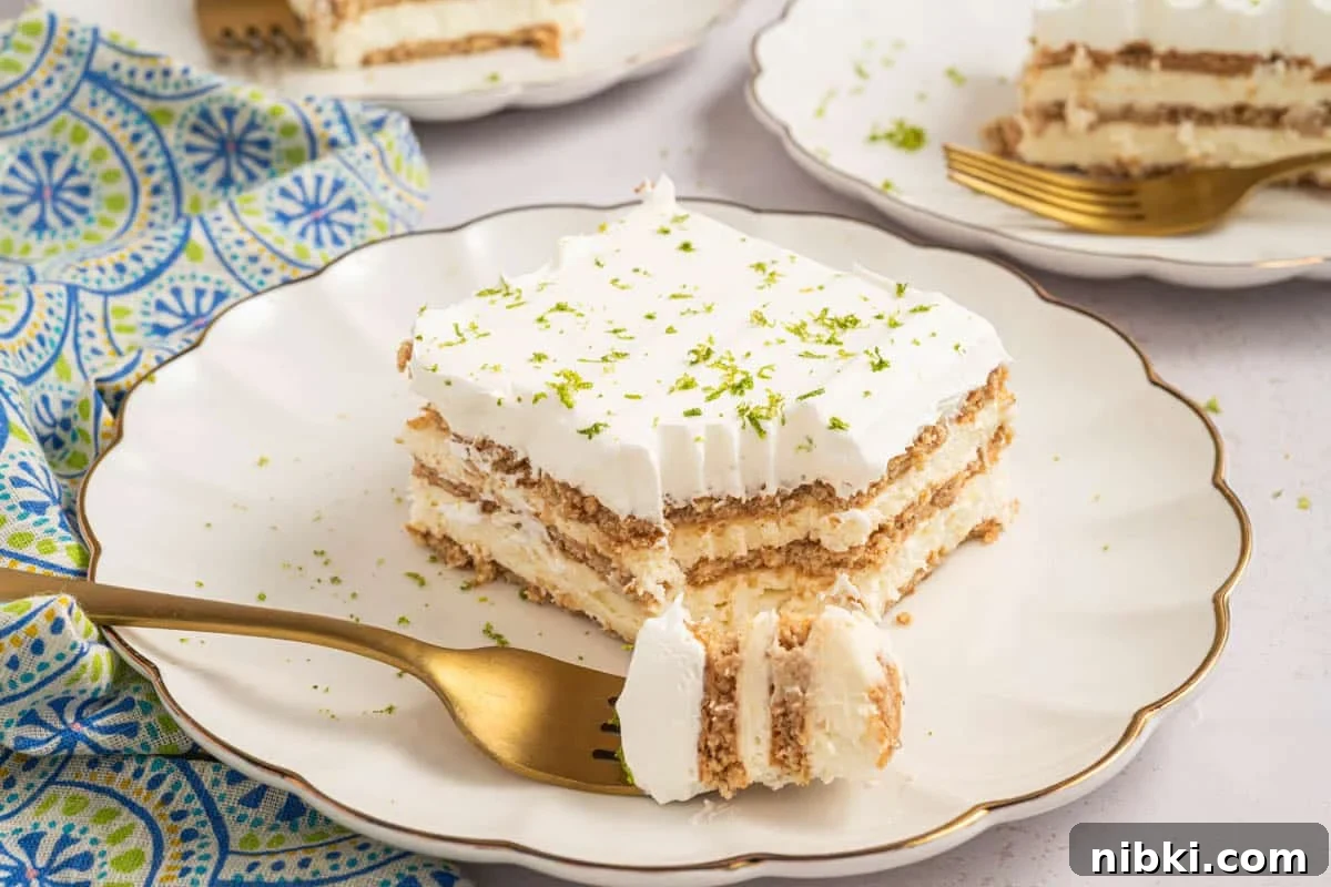 No-Bake Key Lime Icebox Cake Recipe 2 A slice of layered key lime icebox cake with cream and graham crackers on a white plate, topped with lime zest, with a gold fork holding a bite.
