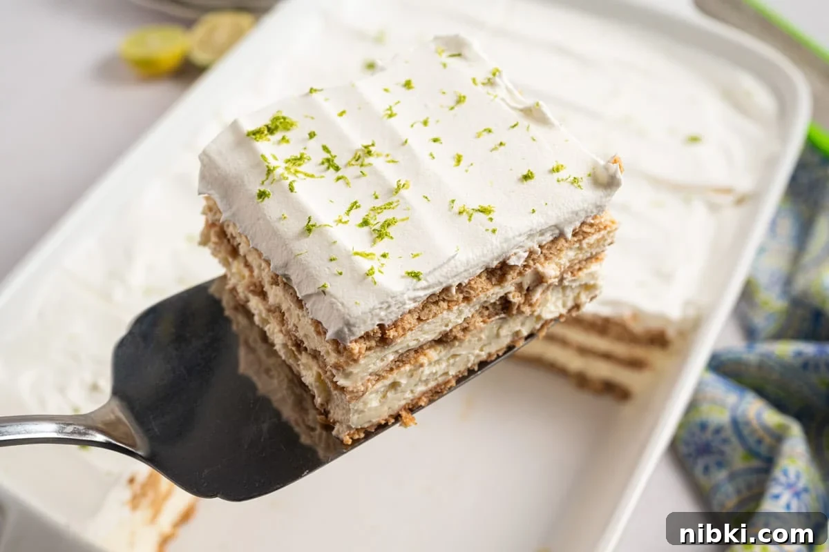 No-Bake Key Lime Icebox Cake Recipe 6 A spatula holds a square slice of key lime icebox cake with whipped topping and lime zest, above a white baking dish containing more of the dessert.