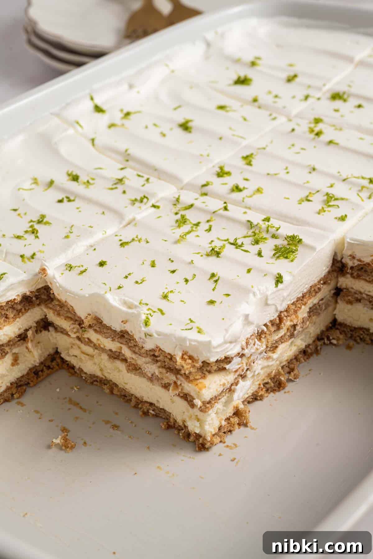 No-Bake Key Lime Icebox Cake Recipe 7 A rectangular dish of key lime icebox cake with whipped topping, graham crackers, and lime zest, with several pieces already served.