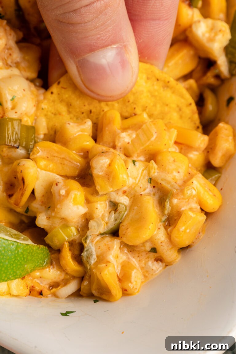 Creamy Mexican Street Corn Dip Recipe 5 A close-up of tortilla chips being dipped into warm street corn dip with corn kernels, cheese, jalapeño pieces, and lime visible in the dish.