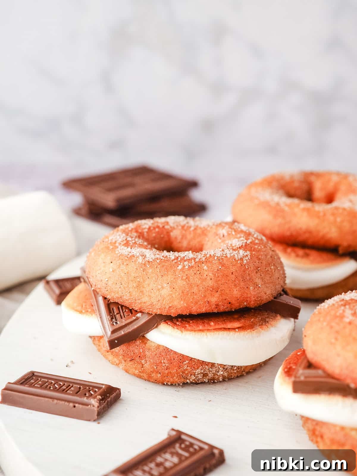 S'mores Donuts Recipe: Chocolate, Graham and Toasted Marshmallow 8 Smore donuts on a serving board with pieces of Hershey’s chocolate and marshmallows.