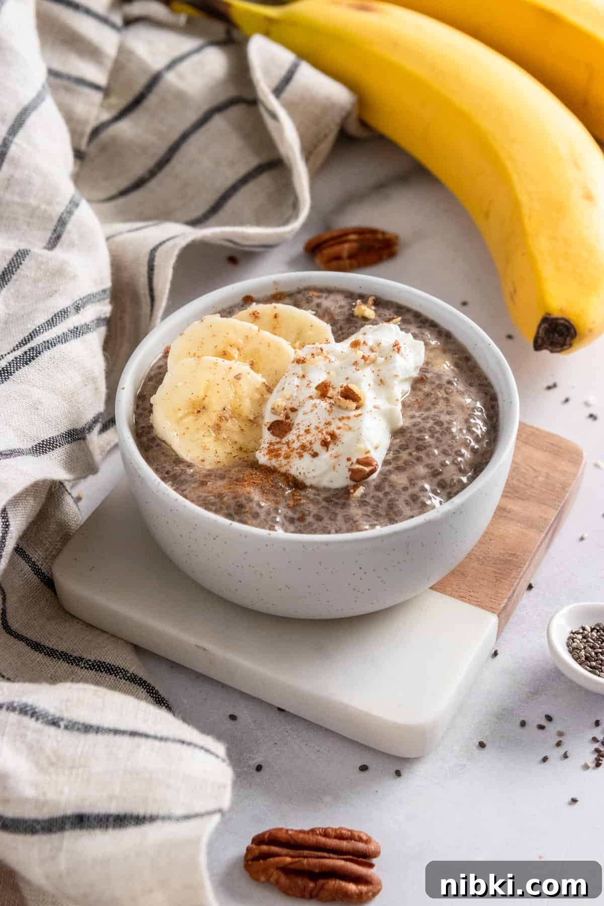 Chia pudding in bowl on coaster with bananas and pecans in the background.