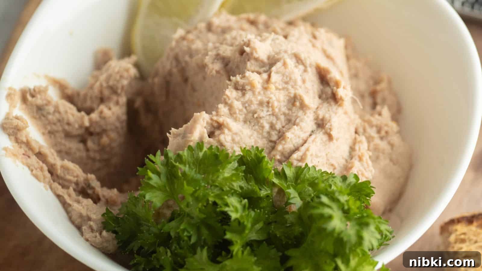 33 Christmas Potluck Recipes to Bring December Magic 2 A white bowl filled with a creamy, light brown spread, garnished with fresh parsley and lemon slices.