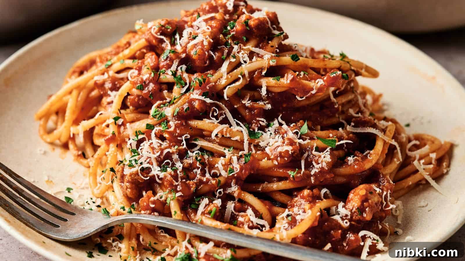 33 Christmas Potluck Recipes to Bring December Magic 32 A plate of spaghetti topped with meat sauce, grated cheese, and chopped herbs, with a fork resting on the plate.
