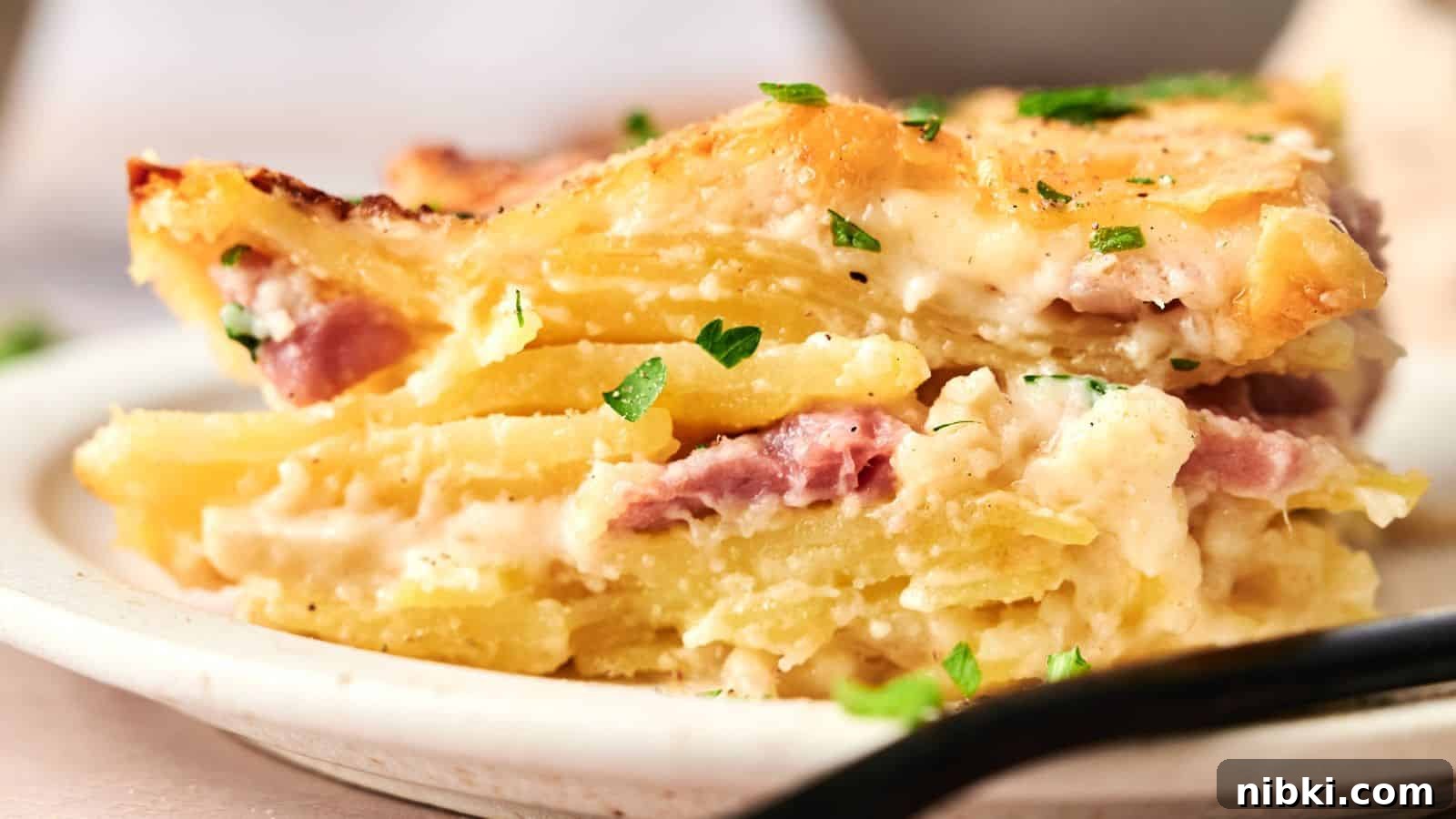 33 Christmas Potluck Recipes to Bring December Magic 7 A close-up of a slice of potato casserole with layers of potatoes, ham, and creamy sauce, garnished with chopped parsley on a plate.