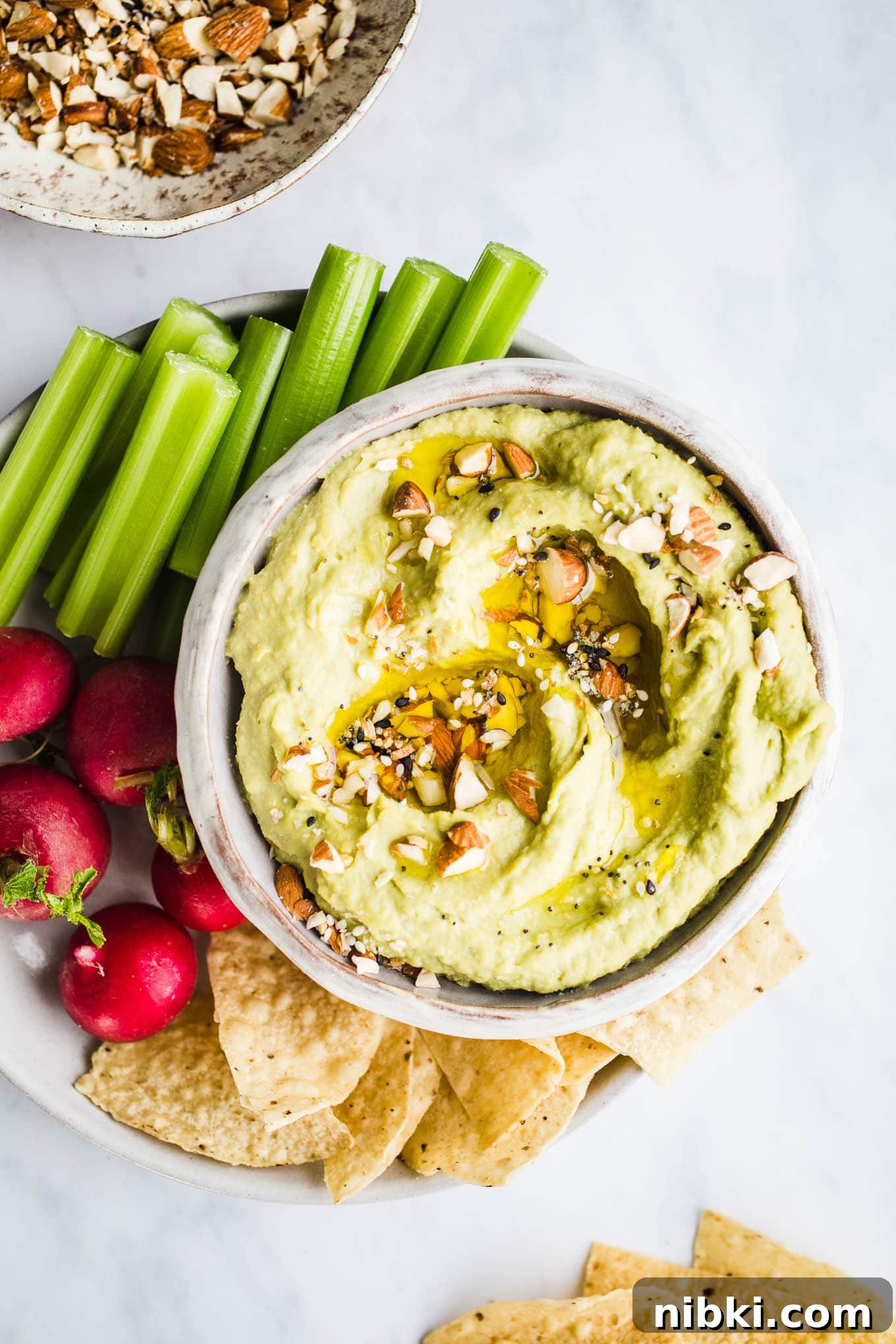 Green avocado hummus in a white bowl served with celery, radishes, and tortilla chips.