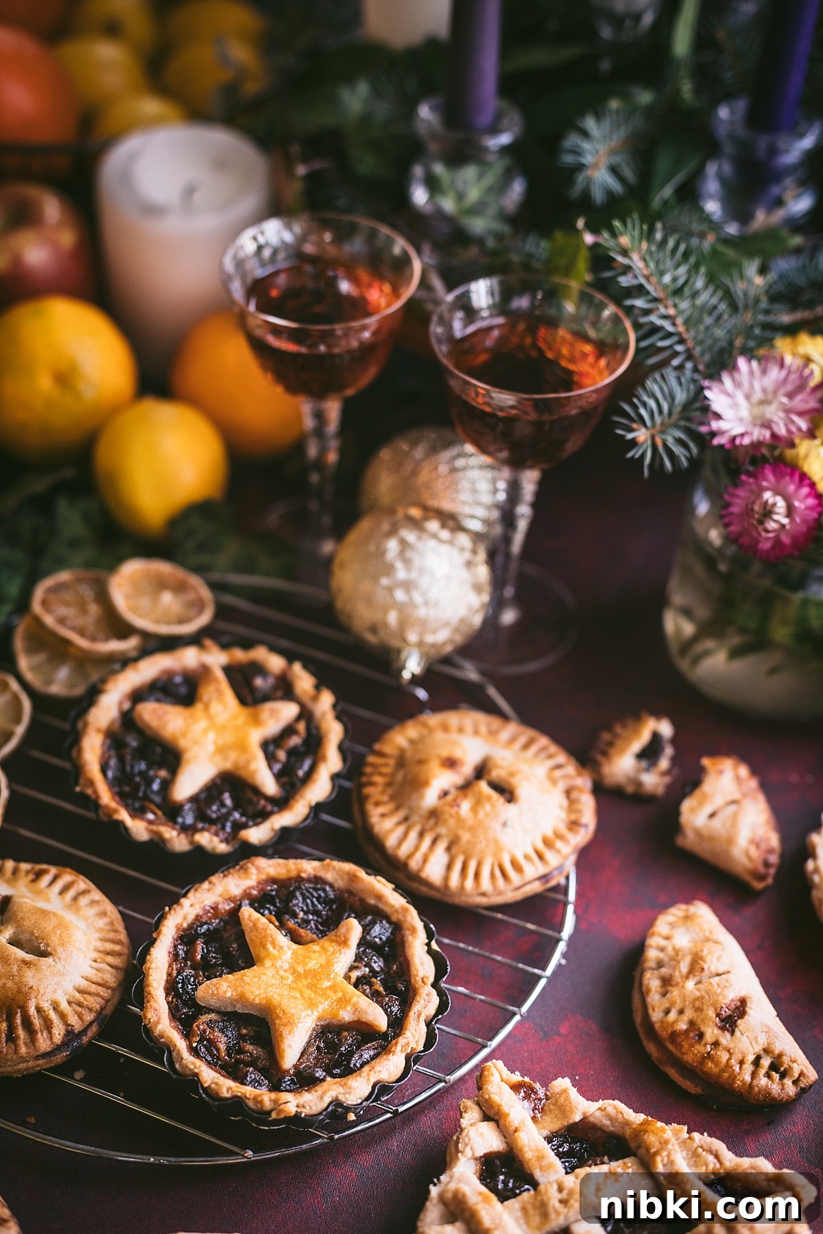 backlit angled shot of two mincemeat pies on cooling rack decorated with star tops. surrounded by grand marnier, greenery, candles, citrus, and apples.