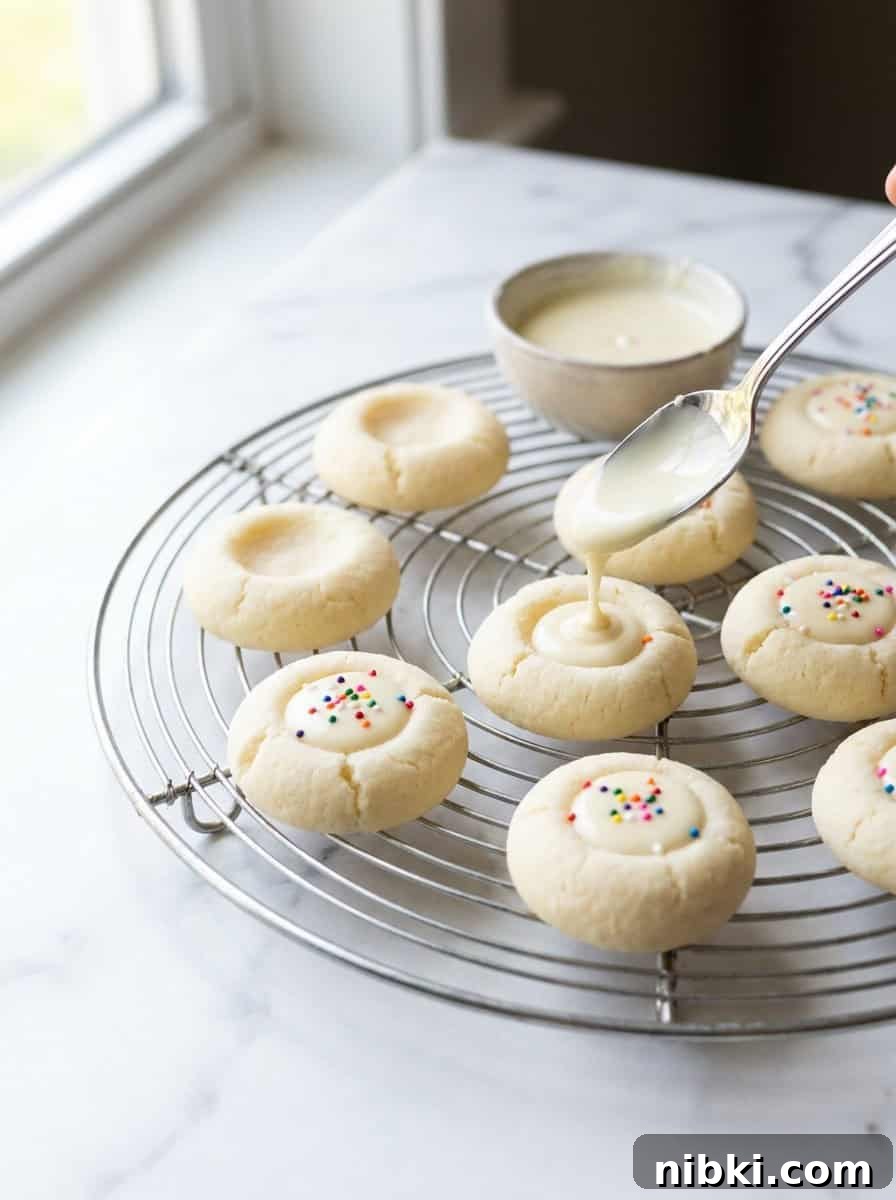White Chocolate Thumbprint Cookies Recipe with Raspberry Jam 3 White cream cheese Thumbprint Cookies on a cooling rack being filled with white chocolate ganache and sprinkles.