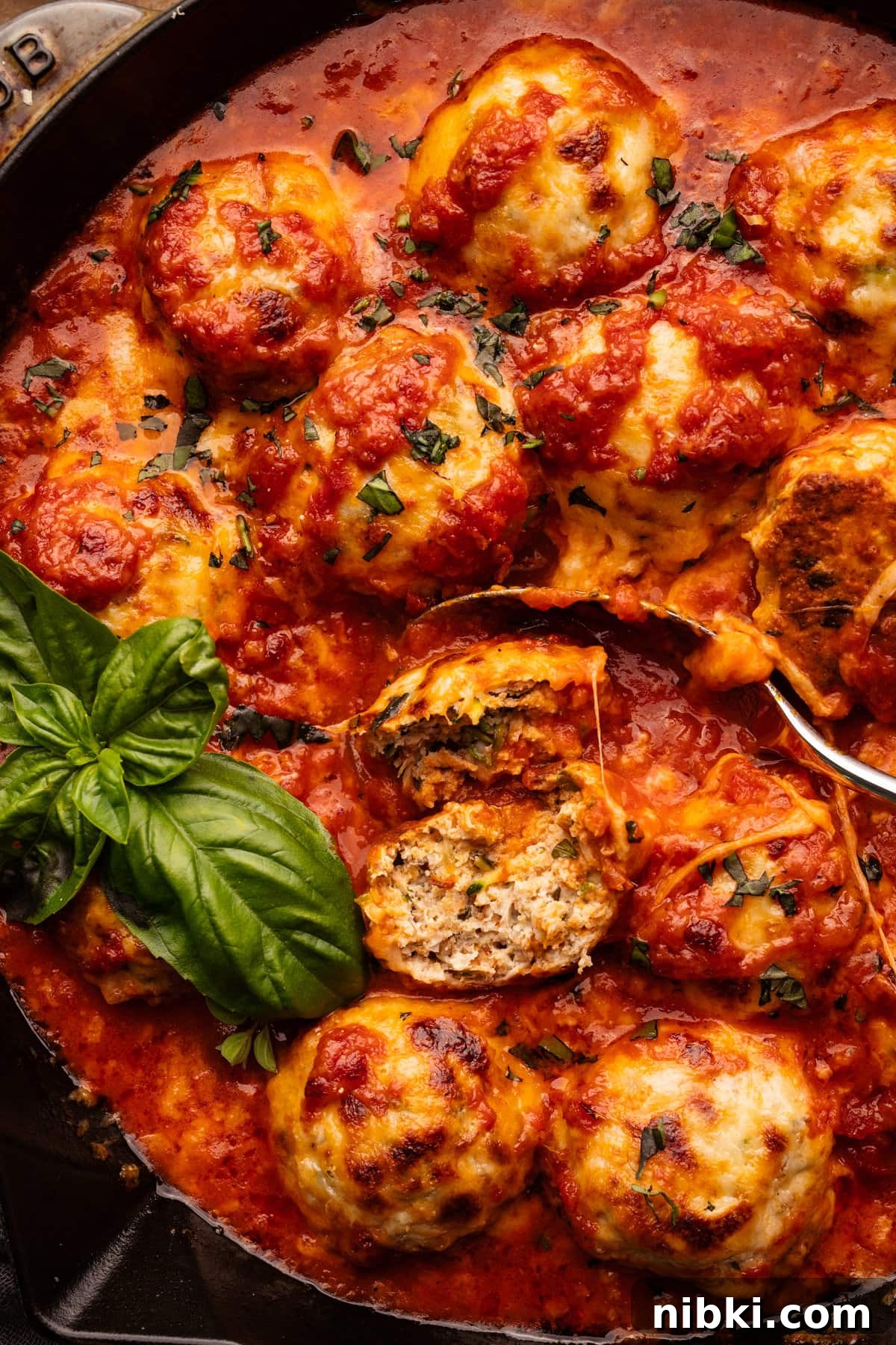 Chicken Parmesan Meatballs with Sauteed Zucchini 5 A close up of chicken parmesan meatballs, one of the meatballs is cut in half with a spoon and fresh basil next to it.