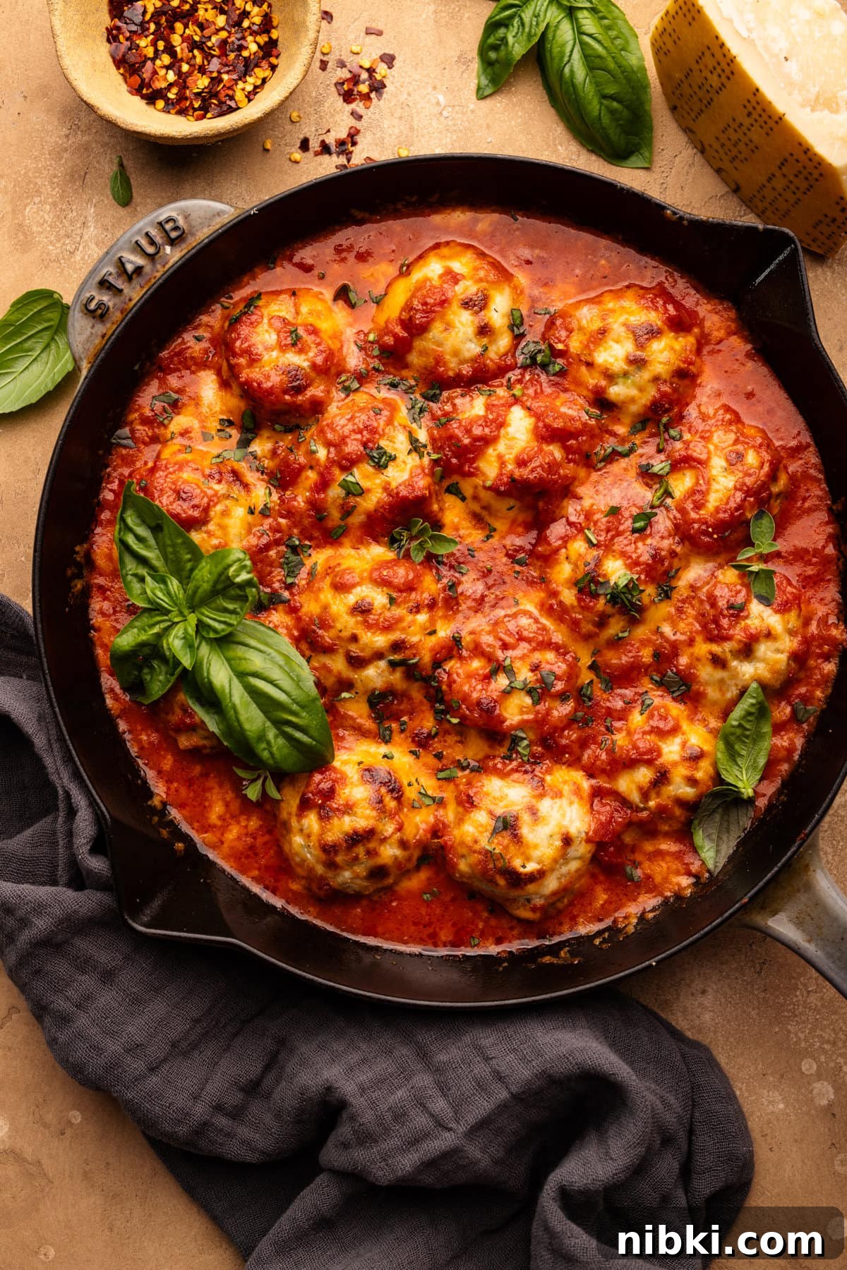 Chicken Parmesan Meatballs with Sauteed Zucchini 6 A skillet pan with chicken parmesan meatballs baked in it with fresh basil on top. The pan is sitting on a tan background with basil leaves, a block of cheese, and a small bowl arranged around it.
