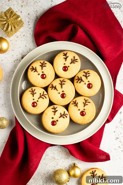 A plate of Reindeer cookies set on a second plate and a red linen napkin. Set on an off white backdrop with golden baubles, golden presents and more reindeer cookies in the background.