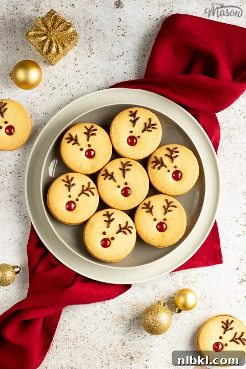 A plate of Reindeer cookies set on a second plate and a red linen napkin. Set on an off white backdrop with golden baubles, golden presents and more reindeer cookies in the background.