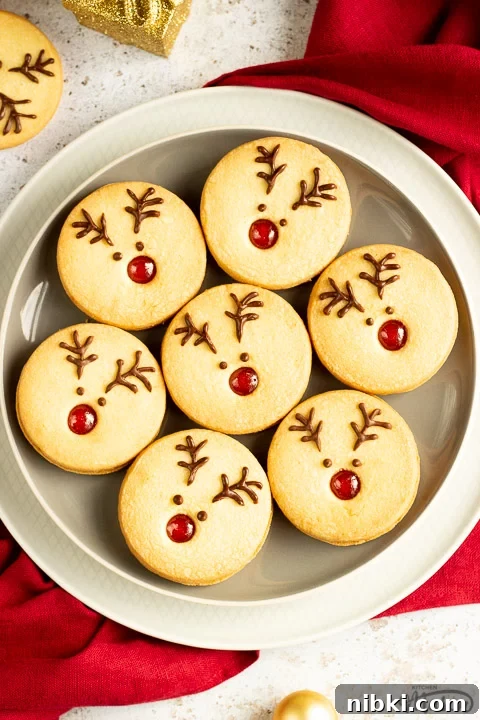 A plate of Reindeer cookies set on a second plate and a red linen napkin. Set on an off white backdrop with golden baubles, golden presents and more reindeer cookies in the background.