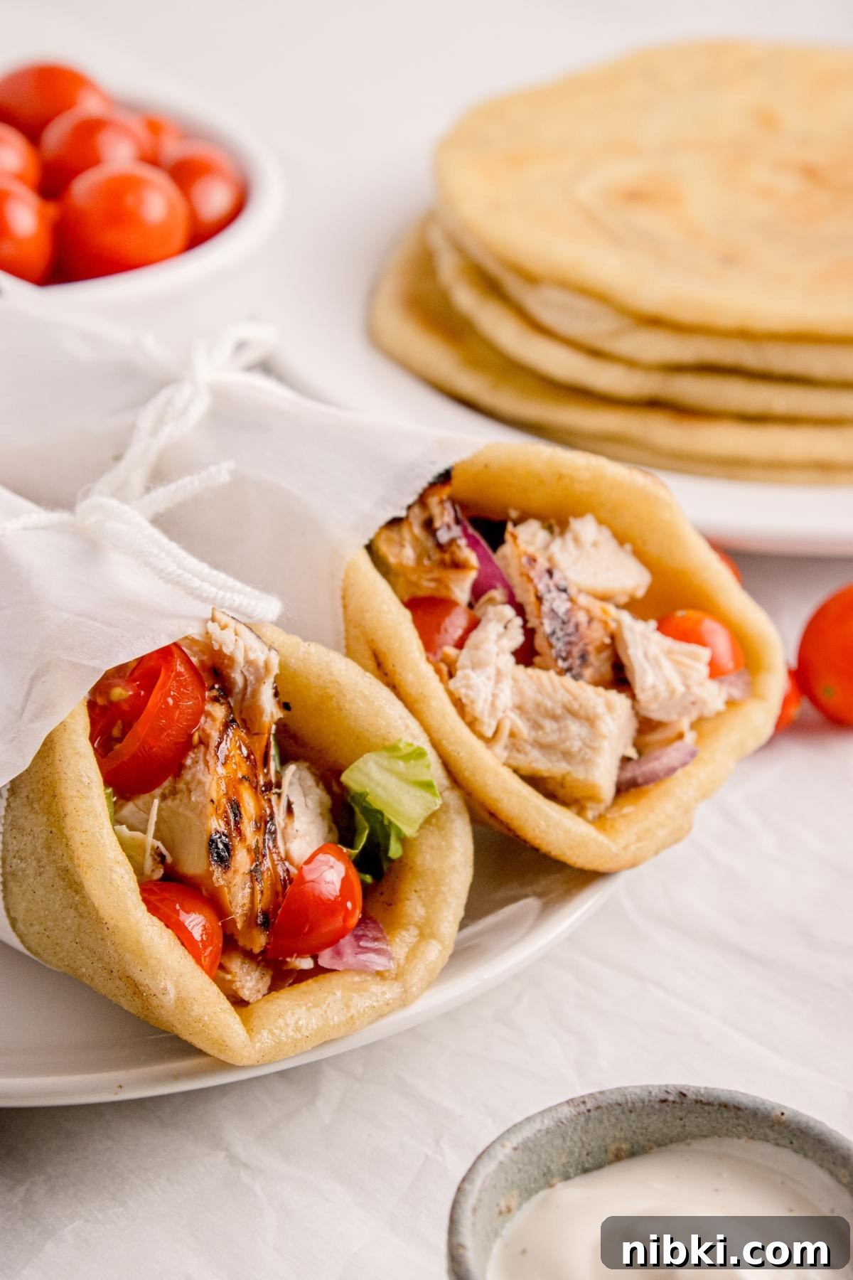 Two pita wraps filled with grilled chicken, lettuce, tomatoes, and onions, with flatbreads and tomatoes in background.