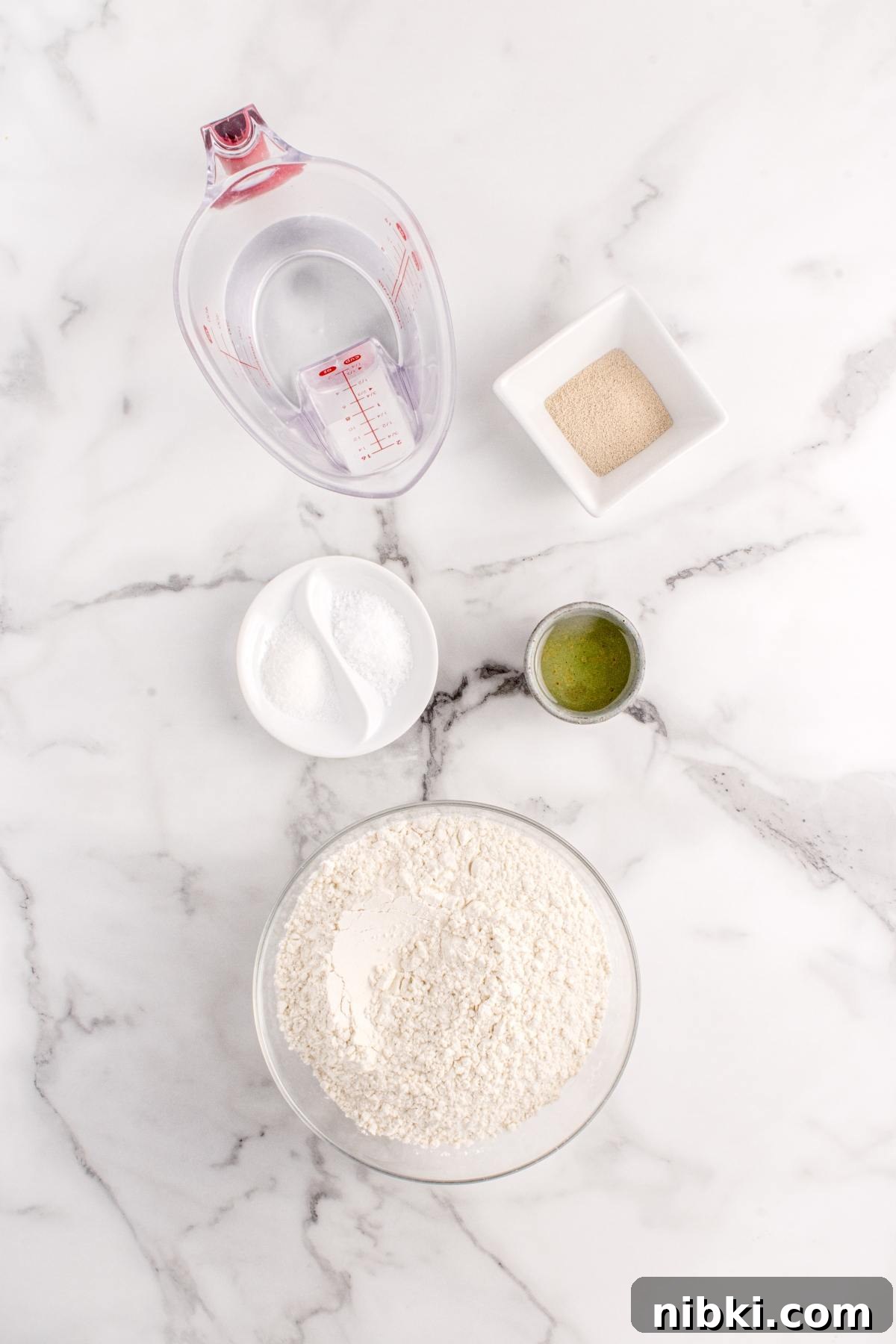 Overhead view of flour, yeast, sugar, oil, and water in separate containers on a marble surface.