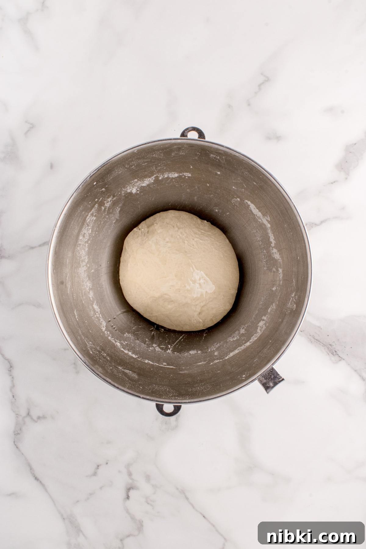 A ball of dough rests in a metal mixing bowl on a white marble surface.