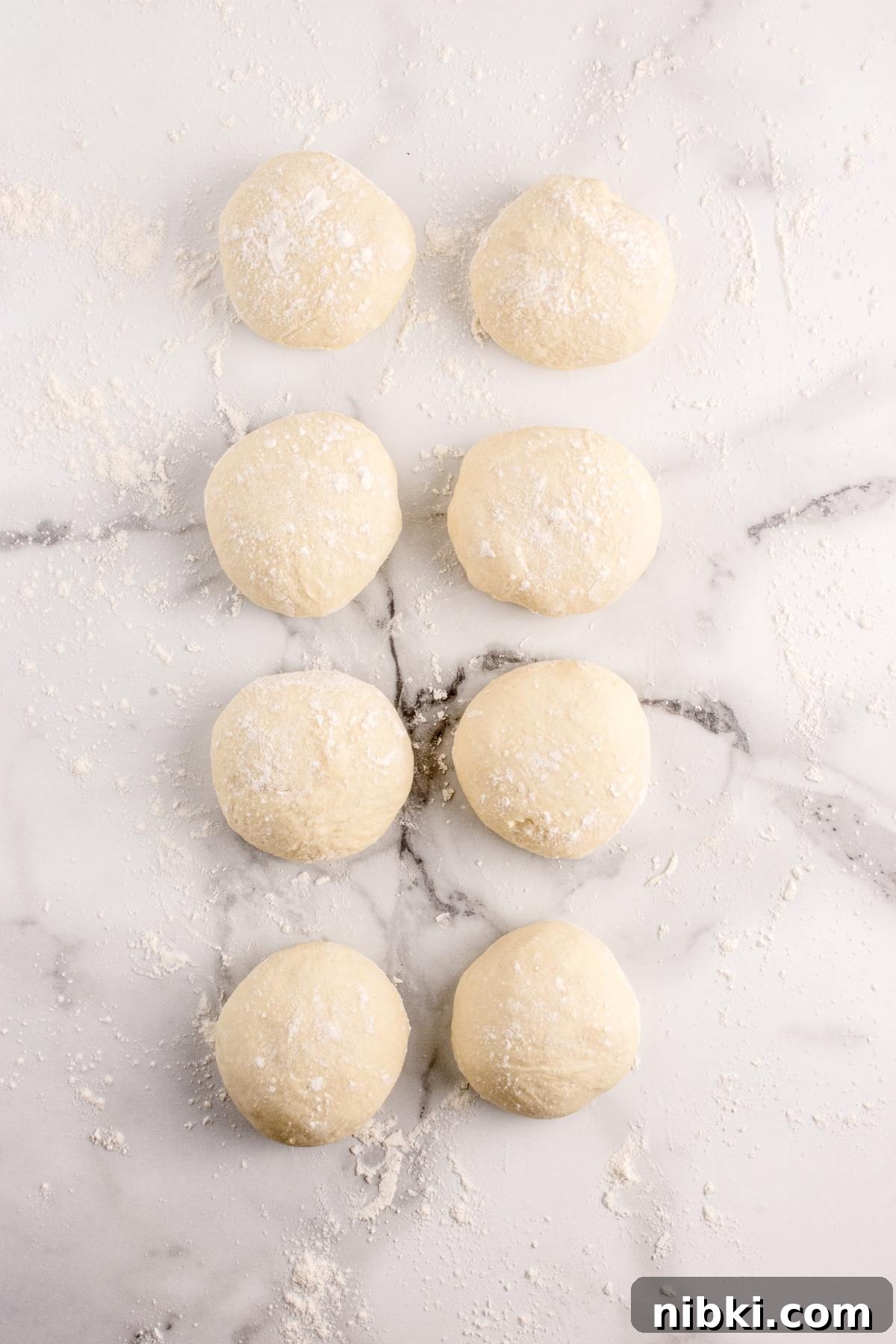 Eight balls of dough dusted with flour are arranged in two vertical rows on a marble surface.
