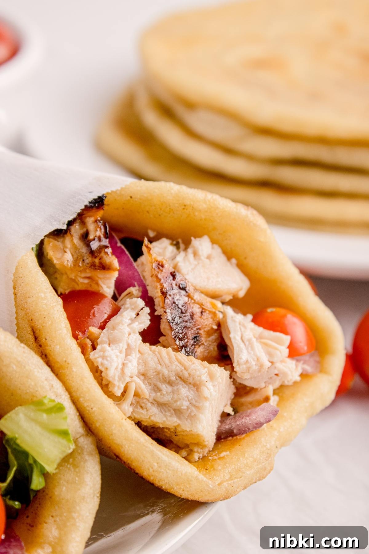 Close-up of a pita stuffed with grilled chicken, tomatoes, and onions, with more pitas stacked in the background.