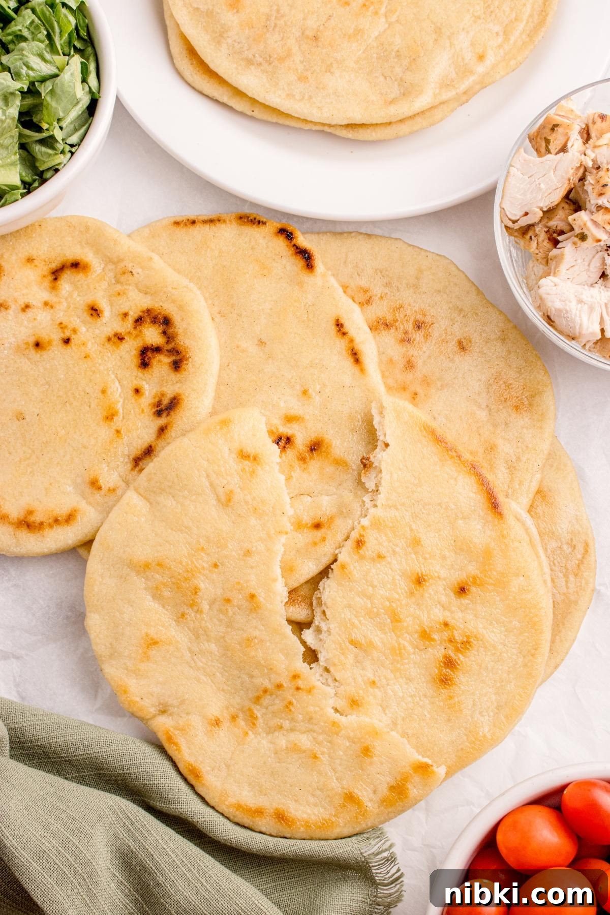 Four pieces of flatbread on a table, surrounded by bowls of greens, chicken, and cherry tomatoes.