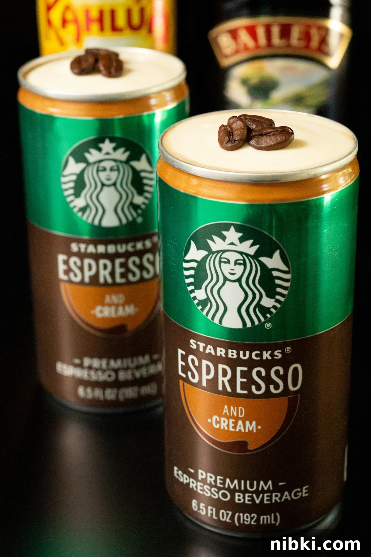 Canned Espresso Martini: Ready-to-Drink Cocktail 3 In focus in the foreground is a Starbucks Espresso and cream can. The top of the can has been removed to show a cold foam topping with three espresso beans sitting on top. Another can is out of focus in the background along with a bottle of Kahlua and Bailey's.