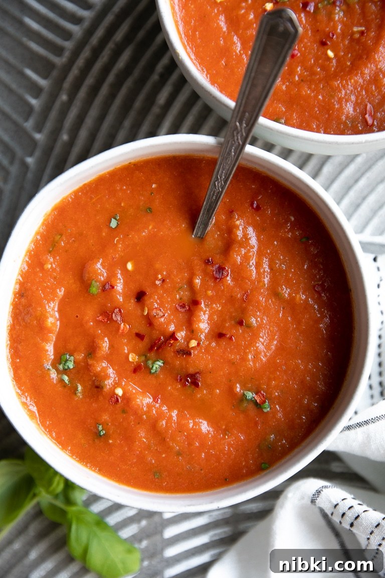 Image of two white bowls filled with homemade tomato basil soup garnished with red chili flakes and fresh basil.