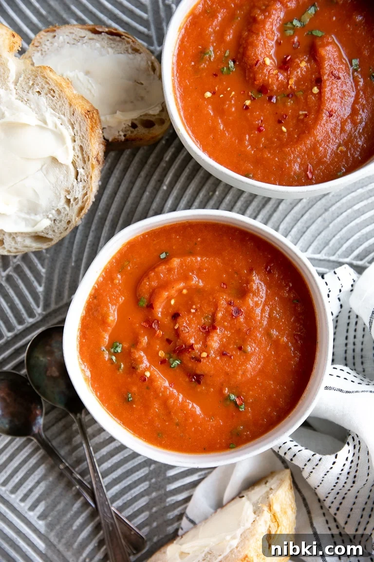 Image of two white bowls filled with homemade tomato basil soup garnished with red chili flakes and fresh basil and served with a side of buttery bread.