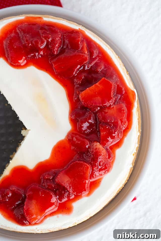 Classic Strawberry Cheesecake Recipe with Fresh Strawberries 2 Top view of a Strawberry Cheesecake with a missing slice, topped with strawberry sauce and pieces of strawberries on a white surface.