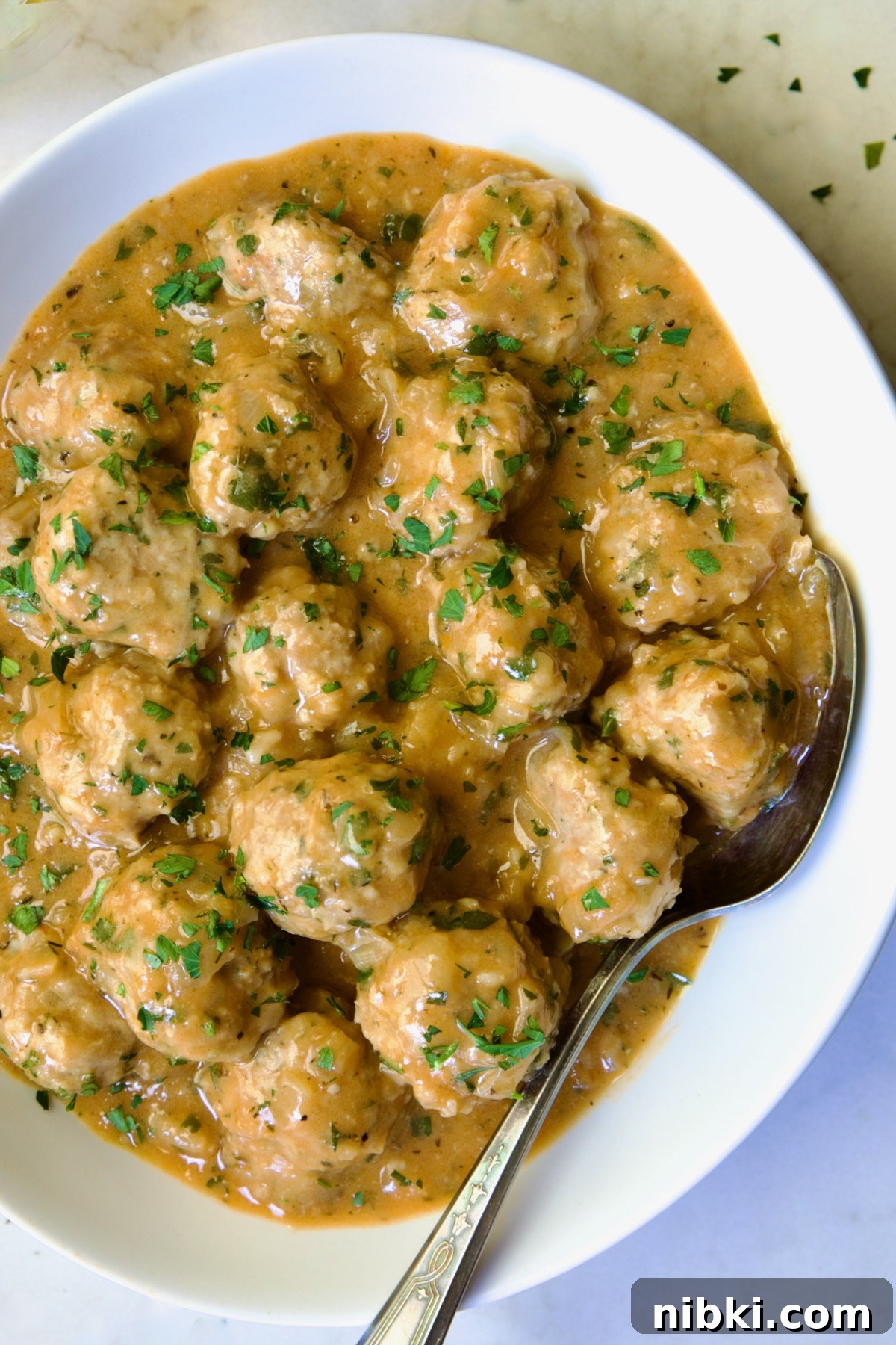 Oven-Baked Turkey Meatballs with Beer Glaze 15 Baked Turkey Meatballs in Beer - Onion Sauce in white oval bowl with serving spoon garnished with fresh parsley.