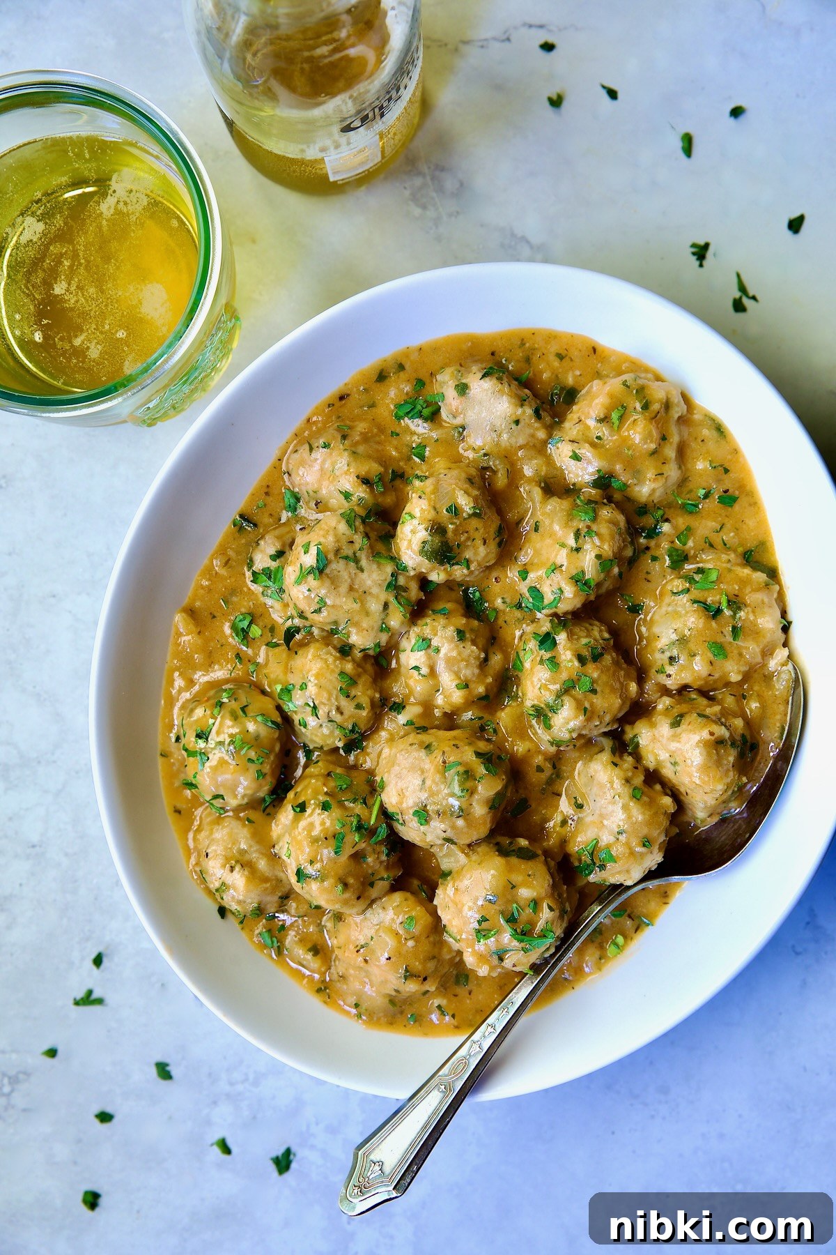 Oven-Baked Turkey Meatballs with Beer Glaze 16 Baked Turkey Meatballs in Beer - Onion Sauce in white oval bowl with serving spoon garnished with fresh parsley.