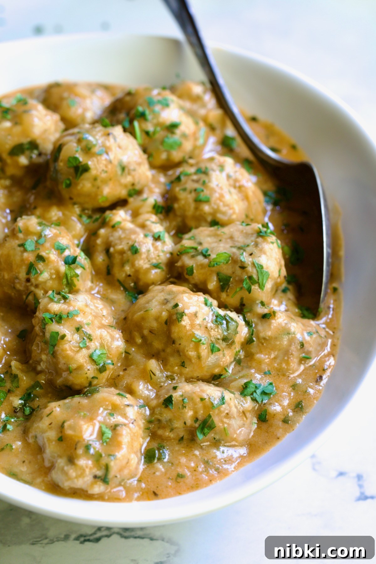 Oven-Baked Turkey Meatballs with Beer Glaze 17 Baked Turkey Meatballs in Beer - Onion Sauce in white oval bowl with serving spoon garnished with fresh parsley.