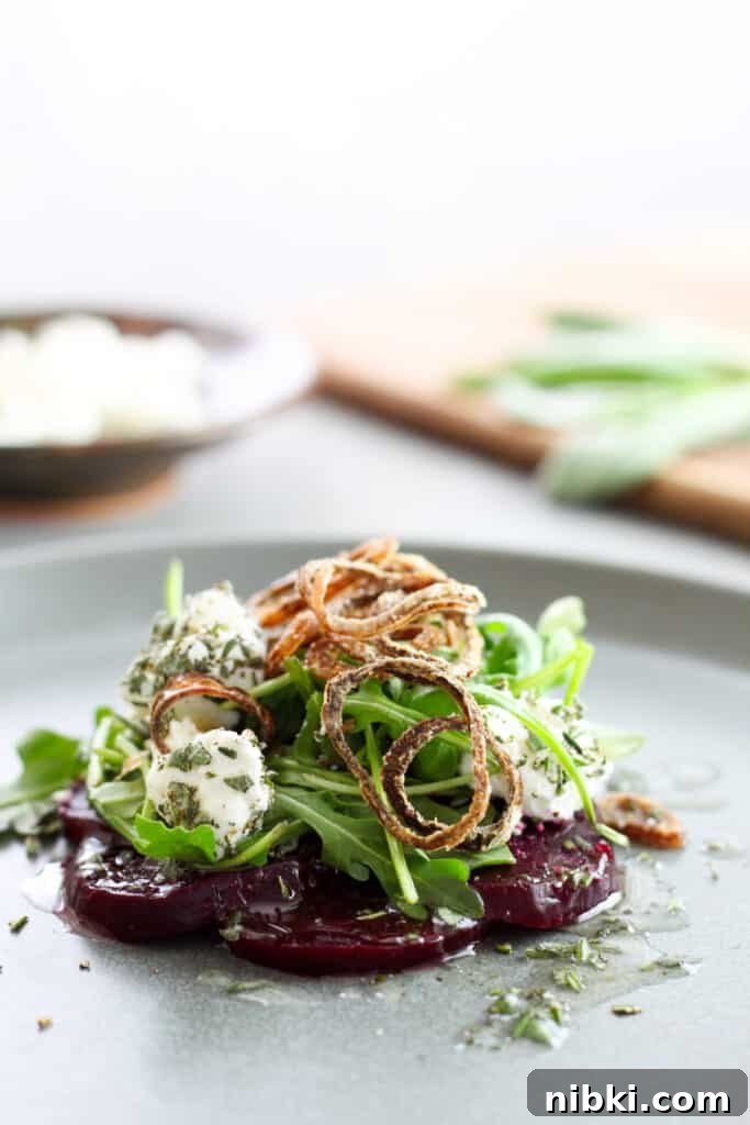 A close up of sliced purple beets topped with arugula, goat cheese rolled in fresh herbs, and topped with crispy fried shallots all on a gray plate. Out of focus in the background is more goat cheese on a small bowl and herbs on a cutting board.