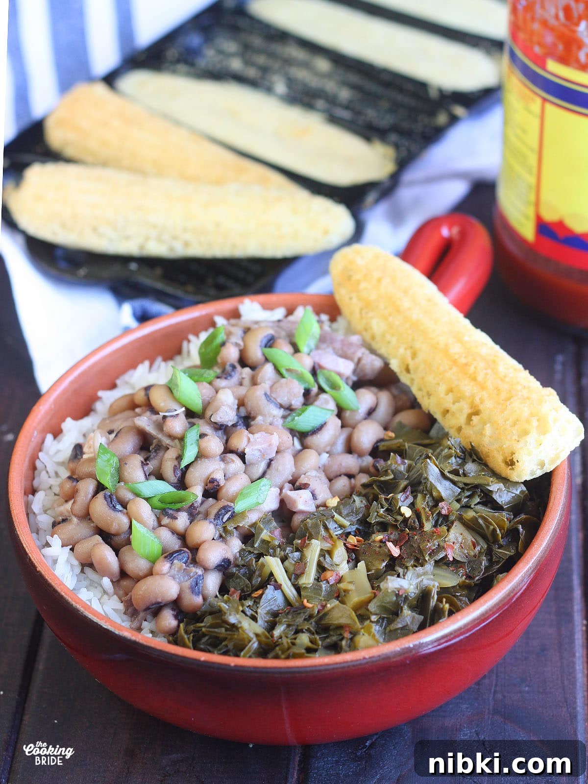 Instant Pot Hoppin' John: Southern Black-Eyed Peas and Rice 9 Red bowl with a handle filled with rice, Hoppin' John and greens. A pan of cornbread and hot sauce is in the background