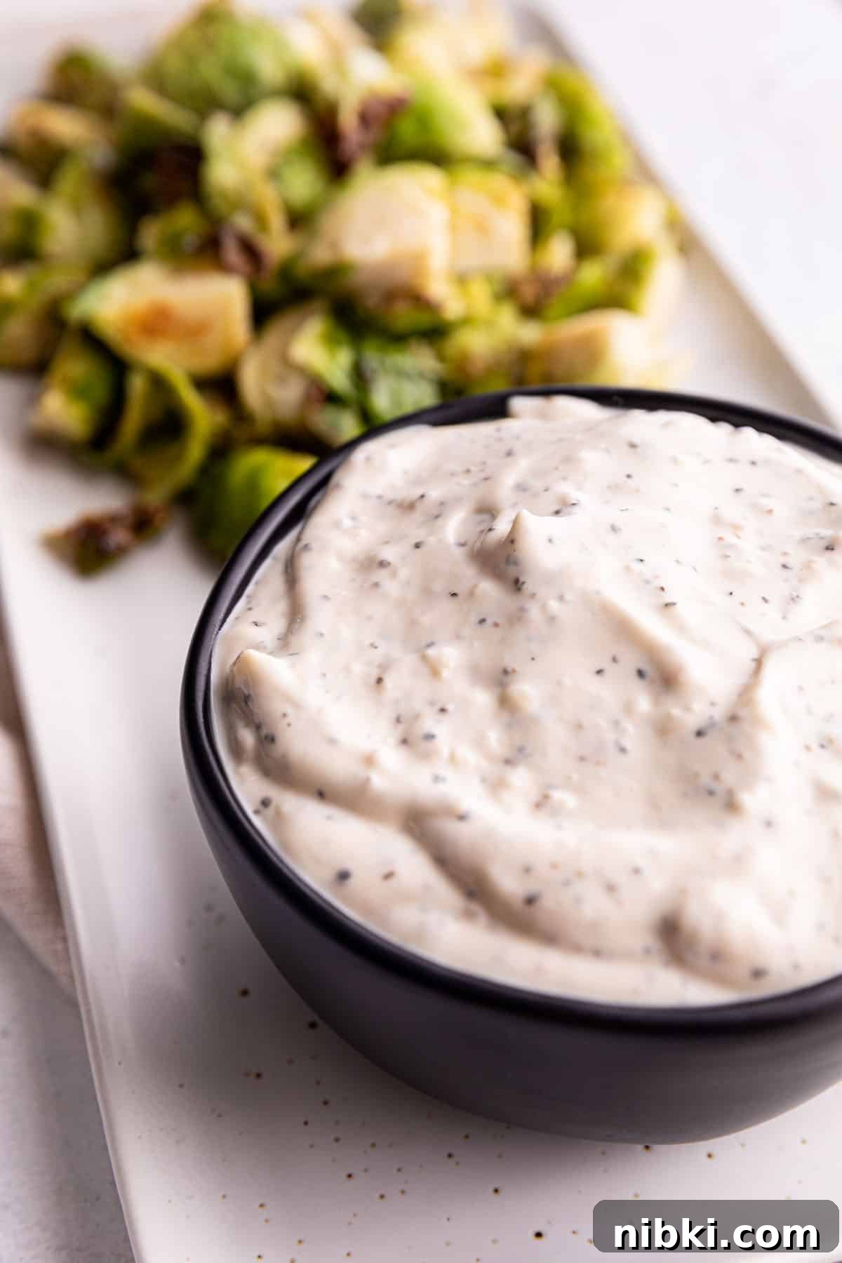 Homemade Lemon Garlic Aioli Recipe 5 A black bowl full of a white sauce with roasted brussels sprouts.